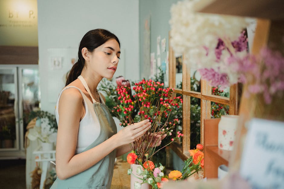 A young woman with dark hair tied back, dressed in a light-colored sleeveless top and beige apron, standing inside a flower shop. She is carefully examining and arranging a bouquet of small, round, pi