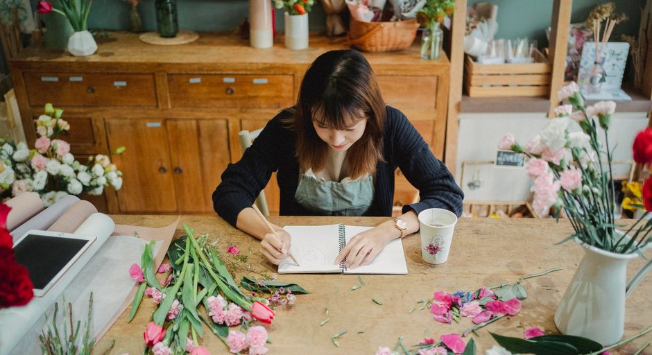 A female florist with shoulder-length brown hair is seated at a wooden worktable inside a flower shop, surrounded by various fresh flowers and floral materials. She is focused on sketching or designin