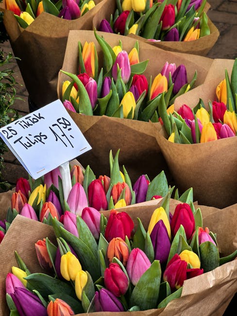 Several bouquets of fresh tulips wrapped in brown kraft paper are arranged on a wooden surface. The tulips display a variety of vibrant colors, including pink, yellow, purple, orange, and red, with gr