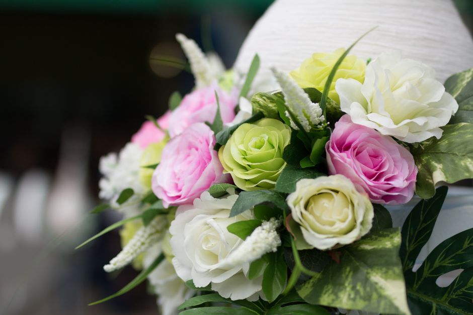 A close-up of a floral arrangement featuring pale pink, white, and light green roses, interspersed with white veronica and lush green foliage. The bouquet is wrapped in elegant white paper and tied wi
