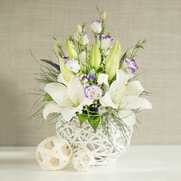 A sophisticated floral centrepiece in white and soft purple tones appears as though it has just been placed on a wedding table at a cosy Stepney venue, bathed in clear, even daylight. The flowers are arranged in a rounded white vase that features an intricate open-weave or rattan-style design, giving it a modern yet natural character, like a sculpted basket rendered in ceramic. Rising gracefully from this textural base, several large white Asiatic lilies open their broad, trumpet-shaped blooms, revealing elegant stamens dusted with pale golden pollen and petals that curl gently at the tips. Intertwined with the lilies are stems of white eustoma, or lisianthus, presenting a range of forms from tightly furled buds to fully open, ruffled flowers edged with a delicate halo of lilac-purple. These soft colours are punctuated by tiny clusters of deeper purple accent blooms, which may be reminiscent of grape hyacinths, adding just enough depth to draw the eye without breaking the arrangement's serene harmony. Fine feathery foliage, similar to asparagus fern or airy ruscus, mingles with broader green leaves, filling out the design and allowing fronds to tumble lightly over the rim of the vase, creating a relaxed, garden-inspired silhouette. Scattered among the larger elements are minute white florets, echoing the look of baby's breath and lending an ethereal, cloud-like texture. The arrangement sits on a smooth white surface, beside two decorative open-weave spheres in soft beige or natural wood tones, which subtly mirror the vase's pattern. Behind everything, a pale beige wall with a softly striated, linen-like texture serves as a calm backdrop, enhancing the brightness of the white petals and the freshness of the greens. The overall impression is one of quiet sophistication and purity, ideal as a wedding table arrangement for ceremonies in Stepney churches, riverside celebrations near the Thames, or as a soothing focal point in a contemporary East London home.