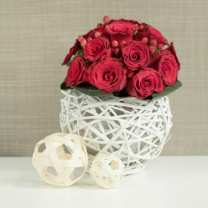 The image showcases a refined, dome-shaped arrangement of red roses designed as a wedding table centerpiece, its form compact and symmetrical yet overflowing with romantic intensity. Dozens of roses in shades of deep crimson and ruby red are tightly clustered, their velvety petals layered in spirals that draw the eye inward. Some blooms are fully open, creating lush, rounded rosettes, while others remain partially furled, adding a sense of life and movement, as if the bouquet is slowly unfolding over the course of an evening celebration. Scattered throughout the roses are small, polished Hypericum berries, their smooth, bead-like surfaces catching the light and bringing a playful, berry-garden texture to the otherwise floral mass. A discreet ring of deep green foliage supports the arrangement from below, visible only at the base, where broad leaves peek out just beneath the blooms and hint at the natural stems and greenery hidden inside. The flowers are gathered into a striking, spherical vase made from finely woven white twigs or branches, arranged in a loose lattice that allows glimpses of the stems and the table beyond. This airy, sculptural vessel rests on a pristine white tabletop, its clean surface reflecting a faint glow from the roses above. To the left, two decorative balls crafted from pale, natural-coloured wicker or rattan lie on the same surface, one slightly larger and closer to the viewer, the other smaller and set further back, reinforcing the organic, textural theme. In the background, a softly blurred wall in warm neutral tones-somewhere between beige and light taupe-adds a gentle, homely context without distracting from the arrangement. The overall composition feels like something created by an experienced Stepney florist for a stylish reception near Limehouse or along the Regent's Canal, ideal for couples seeking a blend of modern design and timeless romance.