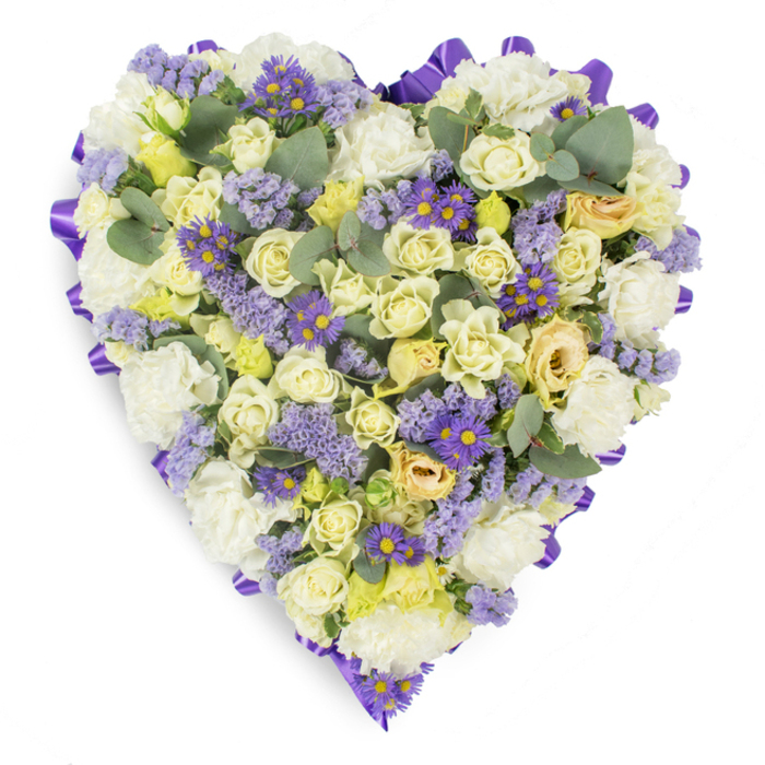 An elegant, heart-shaped floral tribute captured against a pristine white background, where composition and colour work together to convey dignity and solace. The central mass is dominated by cream and pale yellow roses-some tightly furled, others just beginning to breathe-whose layered petals form a sumptuous, textural heart. Interwoven with those roses are generous clusters of white carnations, their ruffled forms adding a soft, classic fullness that evokes tenderness and familiarity. Delicate lavender statice threads through the arrangement, and small, vivid purple daisy-like flowers with prominent yellow centres provide lively, jewel-like accents that prevent the palette from becoming monochrome. Rounded eucalyptus leaves in a cool blue-green are scattered strategically across the surface, their smooth, matte leaves offering both visual rest and a hint of clean, herbal fragrance. A continuous purple satin ribbon is looped around the edge, creating a defined, refined border that enhances the heart silhouette and signals ceremonial intent. The overall feeling is one of peaceful remembrance-the muted creams and lavenders suggesting calm, while the deeper purples quietly affirm respect and affection. I imagine this piece placed at a service in Stepney, beside a remembrance table near Mile End Park, or at a graveside service along Commercial Road; it has the restraint and warmth families seek when offering condolences. Carefully wired and densely packed for lasting presentation, this heart-shaped design communicates love, memory, and serene sympathy.