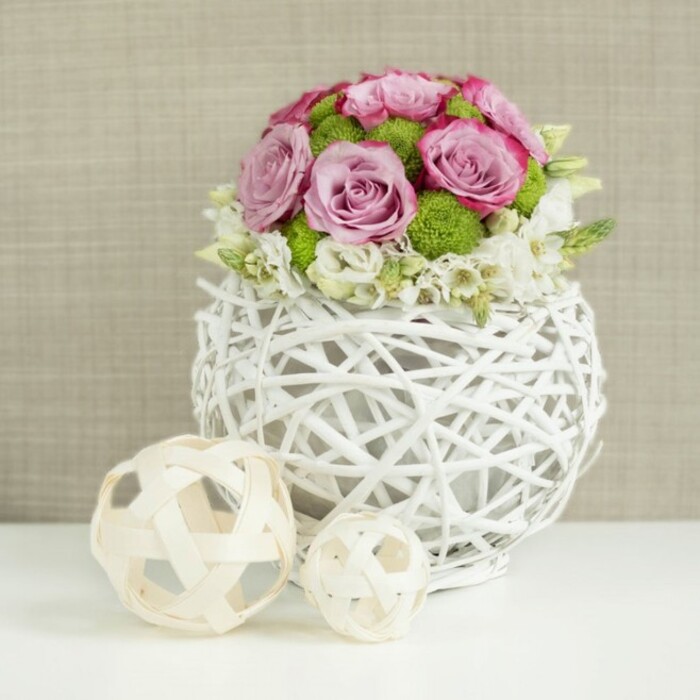 This image shows a beautifully composed wedding table arrangement styled by a florist who understands both modern design and gentle romance, ideal for intimate celebrations across Stepney and the surrounding East London streets. Center stage is a round, openwork vase crafted from slender, twig-like strands painted bright white and woven into a globe shape with visible gaps, so the structure looks light, almost floating above the smooth white tabletop. Filling this airy vessel is a lush, domed bouquet of roses in harmonious shades of blush pink, soft rose, and muted lavender, each bloom fully open and revealing layers of delicate petals edged with subtle hints of deeper pink. Nestled between these roses are small, vivid green button chrysanthemums, their compact, spherical flower heads and velvety texture adding both contrast and a fresh, garden-like feel. At the base of the arrangement and peeking out from between the larger blooms, tiny white or cream filler flowers, reminiscent of baby's breath or small freesias, spill gently over the edge and soften the silhouette, giving the bouquet a cloudlike, romantic finish. To the left of the vase, resting on the same pristine white surface, lie two decorative spheres woven from pale wicker or rattan, one larger and one smaller, echoing the lattice pattern of the vase and enhancing the sense of handcrafted detail. The softly blurred background is a warm beige, textured like linen or light burlap, adding depth without distracting from the florals. Gentle, even lighting washes over the scene, highlighting the curve of each petal and the intricacy of every weave, making the arrangement feel perfect for a wedding reception near Stepney Green, a celebratory dinner in a converted warehouse loft, or a carefully styled photoshoot for local bridal inspiration.