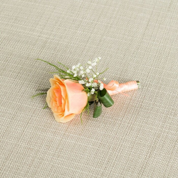 A delicately crafted peach rose boutonniere lies horizontally on a light beige, linen-textured fabric, its refined simplicity capturing the calm moments before a celebration in Stepney begins. The focal rose is partially open, its spiral of petals transitioning from a deeper, warm apricot-peach at the centre to a paler, creamy hue at the outer edges, each petal gently curling with a velvety softness that suggests freshness and a sweet, subtle fragrance. Tucked to the upper right and slightly behind the rose is a soft cluster of white baby's breath, the tiny blossoms forming a lace-like cloud that adds lightness and contrast against the more substantial rose. Wispy, grass-like strands of vivid green foliage emerge from behind the bloom, joined by a pair of broader, darker green leaves that curve protectively around the base, lending structure and a touch of woodland depth, reminiscent of the greenery found along the paths near Stepney Green or the gardens around nearby Limehouse. The stems of the boutonniere are neatly aligned and encased in a smooth peach satin ribbon that closely matches the rose's tones, wrapped in tight, even turns that feel secure and polished, ending in a discreet knot or small bow that anchors the design. The background fabric, with its fine, even weave, adds gentle visual interest while keeping attention firmly on the flowers, and the soft, even lighting casts minimal shadows, highlighting the textures of petal, leaf, and ribbon. This elegant buttonhole suggests a wedding or prom accessory, ideal for a groom, groomsman, or honoured guest attending a ceremony in Stepney, from intimate gatherings in local halls to chic receptions in restored warehouse venues across East London.