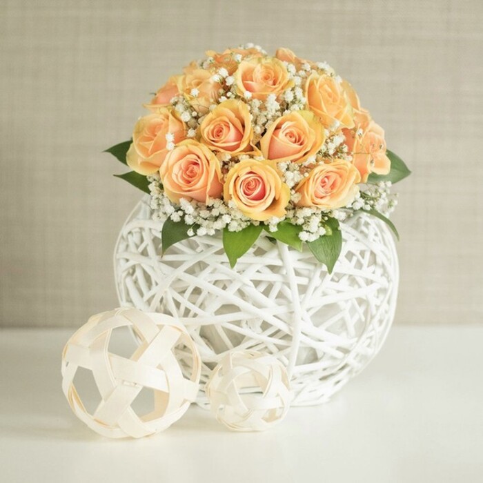 A graceful floral centerpiece composed of soft peach roses and delicate white baby's breath stands on a smooth white table, evoking the quiet elegance of a carefully styled Stepney apartment overlooking Mile End Road. The bouquet is generously rounded, with approximately two dozen roses grouped close together so that their heads almost touch, creating a plush, cushion-like dome. Each rose displays intricate layers of petals, the centres a rich apricot and golden-peach shade that gradually lightens towards the edges, where the colour softens into a creamy, almost pastel tone. This gentle gradient gives the entire arrangement a warm, candlelit quality even in natural daylight. Sprinkled throughout the mass of roses are feathery sprigs of gypsophila, or baby's breath, their tiny white blossoms clustering along slender stems. These add a soft, cloud-like texture that contrasts with the smooth, velvety petals and brings a sense of lightness and romance, much like the subtle floral scent that might drift through a refurbished Victorian home just off Stepney Green. Deep green leaves, partially concealed beneath the blooms, anchor the design with a natural richness and hint at the freshness of flowers recently arranged by hand. The bouquet sits securely in a distinctive spherical vase crafted from white woven material-perhaps wicker or rattan-formed into an open lattice of interlocking bands. This airy structure allows glimpses of shadow and light inside the orb, enhancing the modern, sculptural feel. In front of the vase lie two decorative spheres in a pale beige or natural wood tone, each created from similar woven strips. One is slightly larger, and together they echo the pattern of the container, adding a harmonious, design-led detail. The backdrop is a soft, neutral beige or greige wall with a subtle vertical texture, ensuring the warm roses and intricate weaving remain the unwavering focal point, perfect for a wedding, anniversary centerpiece, or sophisticated home décor in and around Stepney.