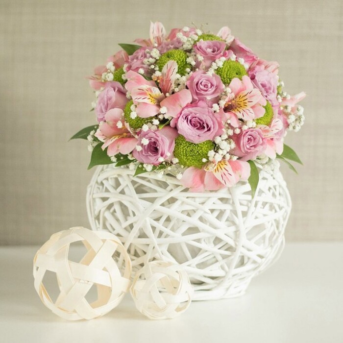 Against a soft, textured beige backdrop reminiscent of Stepney's inviting interiors, a plush and elegant floral centerpiece takes centre stage, radiating warmth and sophisticated harmony. The bouquet features a lush medley of full-bloom pastel pink and lavender roses, their rich, velvety petals unfolding in abundance. Pale pink alstroemeria, each bloom marked by slender petals and the faintest stripes, weave through the arrangement, evoking the subtle artistry seen in local Stepney floral displays. Adding a playful counterpoint, bright lime green button chrysanthemums-loved for their cheery, globe-like forms-bring energy and modern flair. Baby's breath appears in feathery clusters, imparting an ethereal, cloud-like softness that lightens the overall composition. This ensemble is elegantly gathered within a statement white vase distinguished by its intricate, open-weave lattice of slender, intertwined strips. The vase's substantial, round form gives the bouquet both stability and style-a nod to the blend of old and new that characterises the area around Stepney Green Park. Beside the vase, two decorative balls in natural, woven textures-one larger and one smaller-echo the basket-like motif, their placement enhancing balance and cohesion. Set atop a crisp, white surface, this abundant floral display makes a striking yet approachable centerpiece, perfect for wedding tables, sophisticated home décor, or memorable celebrations. The careful mix of luxurious blooms and contemporary accents, thoughtfully arranged by Flowers Stepney, embodies freshness, cheer, and the refined sensibility treasured throughout East London.