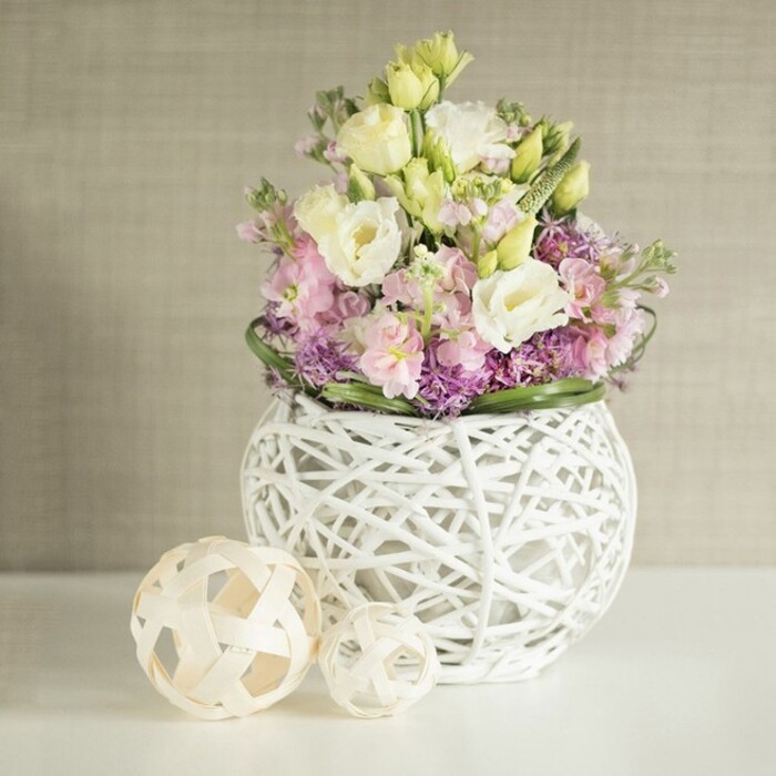 Bathed in gentle, diffused light, a lush floral display sits proudly atop a white table, reflecting the relaxed yet refined atmosphere of a Stepney gathering. The arrangement's focal point is the eustoma-or lisianthus-whose creamy ruffles and pastel yellow tones create a softness mimicking spring's first blooms in local gardens. These are paired with tall, blushing pink matthiola (stock) flowers, their spires adding both height and an air of gentle sophistication. Beneath, clusters of deep purple filler blooms and finely textured asters evoke the wildflower edges found near Mile End's green spaces, while a halo of glossy green foliage brings structure and vivacity. The contemporary vase is crafted from intricately woven white branches or painted rattan strands, forming a spherical, openwork container with an almost sculptural quality, its lattice design casting playful shadows. To the left, two decorative spheres of pale rattan rest easily, echoing the artistry and craftsmanship historically celebrated throughout Stepney's street markets and artisan shops. The entire arrangement is positioned against a soft beige wall, its vertical texture subtly referencing the drapes and wallpapers common in historic East London homes. Together, these elements create an image radiating calm and natural elegance-perfect for a Stepney wedding reception table, modern apartment, or as rich inspiration for local DIY centerpiece creation.