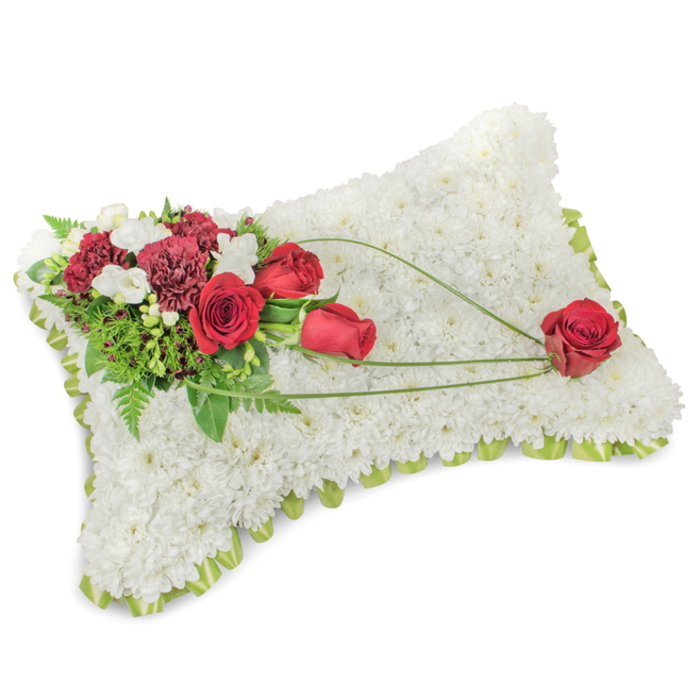 This elegant pillow-shaped memorial arrangement lies against a pure white backdrop, its form defined by a dense carpet of pristine white chrysanthemum blooms. Each small flower is tightly set to produce a tactile, velveteen surface that reads as comforting and pure. A narrow band of pale green-either satin ribbon or fresh foliage-edges the slightly raised cushion, offering a subtle frame and a hint of spring-like freshness. Anchoring the design at the upper left is a lavish spray of deep red roses paired with dark burgundy carnations; the roses' velvety petals show stages of bloom from tight centers to full-open cups, their colour a vivid, respectful contrast to the white ground. Scattered among this cluster are tiny white filler blooms, likely freesias or spray chrysanthemums, which add delicate texture and a whisper of scent. Greenery-feathery ferns and broader leaves-recedes behind the flowers, creating depth, while three elegant arcs of bright green grass-like foliage or decorative stems sweep diagonally across the pillow, guiding the gaze to the lower right. There, a single, perfectly formed red rose rests as a gentle emblem of remembrance, its placement feeling intentional and tender. The photographer's soft lighting enhances the arrangement's textures and calm mood. A local florist in Stepney would choose this classic sympathy cushion for services near Stepney Green, St. Dunstan's, or Tower Hamlets Cemetery, where its quiet beauty and measured craftsmanship convey respect, comfort, and lasting affection.