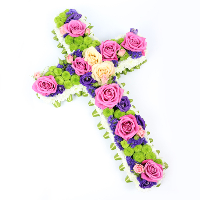 A beautifully composed floral cross dominates the image, set against a bright white background that highlights every nuance of its design. The cross is shown at a slight diagonal, the top left extending gently forward toward the viewer, giving a subtle three-dimensional sense as if it were resting in a quiet Stepney chapel before a service. The outline is defined with precision: small white spray chrysanthemums are massed tightly together to form a plush, snowy border, each tiny bloom adding to the impression of softness and purity. Just beneath this layer, a band of light green ribbon or carefully folded foliage circles the shape, offering a delicate rim of colour that echoes the fresh greenery seen around local Stepney gardens and squares. Inside this graceful frame, the cross is abundantly filled with a lively mix of flowers. Large roses in bright fuchsia pink and softly toned pale yellow or creamy peach are positioned thoughtfully along the length and arms, creating focal points that naturally guide the gaze. Between these roses sit clusters of spherical lime-green button chrysanthemums, like little pom-poms of colour, adding texture and a modern, uplifting twist. Woven throughout the central arrangement are smaller, deep purple and violet blossoms-perhaps lisianthus or statice-nestling between the larger blooms and introducing a richer, darker tone that deepens the overall palette. The lighting is clear and even, revealing the satin sheen of the green ribbon, the velvety surfaces of the rose petals, and the intricate structure of each chrysanthemum. The overall mood is one of quiet dignity and gentle hope, a floral cross ideal for funerals, memorials, or graveside tributes for families in Stepney seeking a meaningful expression of remembrance and faith.