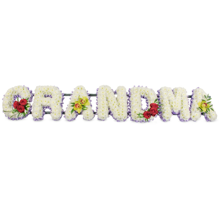 In this image, the word GRANDMA is lovingly spelled out in large floral letters, capturing the feeling of a family tribute prepared for a final farewell in Stepney. The letters are set side by side in a straight row against a crisp white background, allowing every petal and colour nuance to be clearly seen. Each character is entirely filled with small, snowy white chrysanthemums or similar tightly petalled blooms, arranged so densely that the surface appears smooth and softly padded, like a cushion of flowers. Beneath this blanket of white, a fine edging of light lavender or pale purple blossoms peeks out along the bottom of each letter, adding a gentle trace of colour that defines the outlines without overpowering the calm, pure palette. On the graceful curve of the opening G, three deep red roses rest at the lower left, their rich tones and layered petals symbolising deep affection and remembrance, supported by dark green leaves that provide contrast and natural movement. The first A introduces a pair of yellow-green cymbidium orchids, carefully placed across the crossbar, their fresh, chartreuse petals and subtle reddish-brown centres offering a hint of modern elegance. The central R, N, and D are kept in simple white and lavender, drawing attention to the craftsmanship and clarity of the lettering itself. On the M, another grouping of three red roses nestles into the lower right arch, their colour echoing the G and creating a sense of rhythm along the tribute. The final A repeats two yellow-green orchids, balancing the design from end to end. All letters are secured to a slim, dark green or black framework that remains mostly hidden, giving the impression that the flowers float together as one unified piece. The arrangement conveys serenity, dignity, and familial love, well suited for funerals, wakes, or graveside gatherings throughout Stepney, Limehouse, and the surrounding East London community.