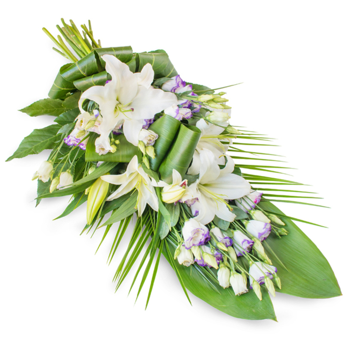 An elegant, elongated sympathy spray is shown against a bright white background, its classic teardrop shape reminiscent of the graceful floral tributes often seen at memorials across Stepney and the wider East End. The composition begins with a generous cluster of stems at the upper left, then gently flows downward to a narrow, tapered point at the bottom right, creating a sense of direction and quiet movement, as though the flowers were bowing in a final gesture of honour. At the centre of this design are several striking white oriental lilies, some fully unfurled with wide, sculpted petals and softly tinted stamens, others still enclosed in smooth, tapering buds. They are surrounded by numerous white lisianthus (Eustoma) flowers, whose soft, ruffled petals carry the faintest hint of lavender or pale purple along their edges and veins, bringing subtle colour and a sense of delicate refinement. The floral elements are cradled in a lush frame of greenery. Deep green, broad leaves form a solid, reassuring base, evoking the mature trees around Stepney Green, while long, slender palm-like fronds sweep gracefully out to the right, accentuating the overall line of the spray. Some leaves are artfully folded and rolled into sculptural forms near the centre, showcasing the florist's craft and adding visual interest without overwhelming the serene mood. The balanced interplay of creamy whites, gentle purples, and rich greens creates an atmosphere of peace and dignity. This refined funeral arrangement would be fitting atop a casket, beside a memorial photograph, or at a graveside service, conveying compassion, remembrance, and respect to families gathering in Stepney to say their goodbyes.