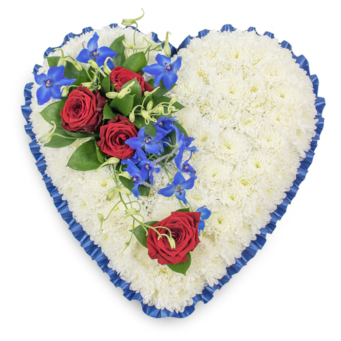 A beautifully composed, heart-shaped funeral tribute is shown from above, its outline strong and clear against a simple, pale background. The central surface of the heart is entirely covered with compact white chrysanthemums, each flower head carefully placed so that no gaps show, creating the impression of a soft, quilted cushion. The white blooms offer a feeling of calm and purity, their small petals forming layers of texture that read as both gentle and precise. Framing the heart, a royal blue satin ribbon is tightly pleated, the ruffled edge catching the light and lending a subtle sheen that adds formality and grace. On the upper left side, a striking floral spray rests on top of the chrysanthemum base: three fully open red roses, rich and velvety, are cradled in dark green foliage, their deep hue symbolising lasting love and respect. From around these roses emerge slender stems of electric blue dendrobium orchids, each blossom star-shaped and luminous, some blooms curling softly down towards the centre of the heart. Delicate light green tendrils weave naturally through the orchids and foliage, softening the structure and suggesting gentle movement. Balanced on the lower right curve, a single red rose lies on the white flowers, echoing the main cluster and adding a quiet, personal note of remembrance. The overall design feels thoughtful and composed, the kind of tribute that could be delivered by a local Stepney florist for a funeral service near Mile End Road or a memorial gathering at a nearby chapel. It conveys sympathy and love in a refined, dignified way, using colour and texture to speak when words are difficult.