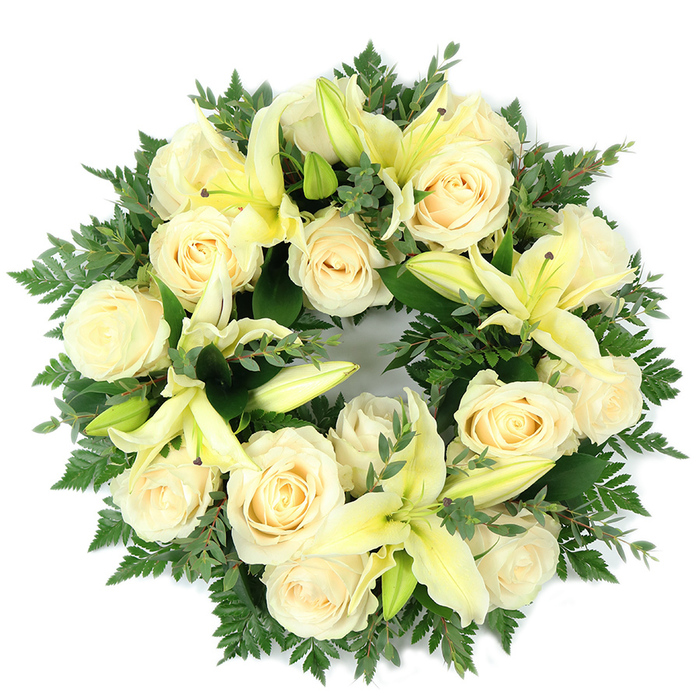 A meticulously arranged, circular sympathy wreath photographed flat against a clean white surface, where creamy white roses and soft yellow lilies are set into a dense, verdant foundation of mixed foliage. Approximately a dozen to fifteen fully opened roses, their petals tightly furled in classic spirals, create the main visual field; pale lemon-hued lilies, with some blossoms fully unfurled and others held as graceful buds, rise through and above the roses, lending subtle height and elegant trumpet-shaped lines. The contrast between the warm, gentle tones of the blooms and the cool, varied greens is deliberate: fine, feathery fern fronds soften the wreath's silhouette while sprays of broader, rounded leaves-likely eucalyptus or a similar species-add silvery-green shadows and texture. Soft daylight from above caresses the petals, producing a calm interplay of highlights and shadow that emphasises each flower's form and the overall round geometry. The wreath's full, luxurious packing reads as both respectful and composed, a visual language of purity, peace and sustained remembrance suited for services in Stepney and the surrounding East End neighbourhoods such as Bow and Bethnal Green. There is an implied, gentle fragrance-the roses' sweet powdery note tempered by the lilies' green-citrus freshness-that deepens the sensory impression and the sense of crafted care. Balanced, symmetrical and dignified, the arrangement stands as a composed tribute for a funeral, memorial table or graveside commemoration where quiet beauty and heartfelt condolence are required.